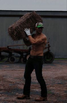 a shirtless man holding a large pile of hay in front of a barn building