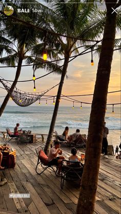 people sitting at tables on a wooden deck overlooking the ocean with hammocks hanging from palm trees