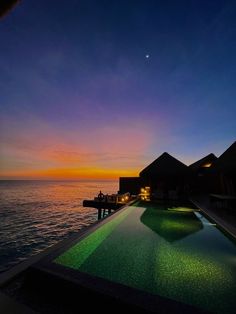 an outdoor swimming pool at dusk with the sun setting in the distance and some houses on the water