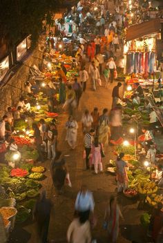 many people are walking around an outdoor market at night with lights on the tables and in front of them