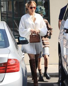 a woman in white shirt and skirt carrying a brown purse walking down the street next to cars