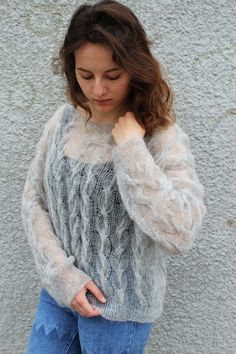 a woman standing in front of a wall wearing a gray sweater and blue ripped jeans