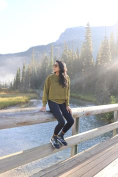 a woman standing on a wooden bridge next to a body of water with trees in the background