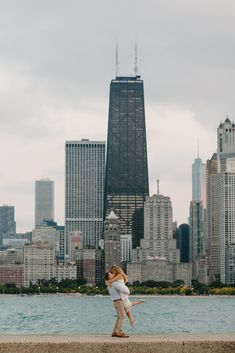 a woman is standing on the edge of a wall by the water in front of a large city