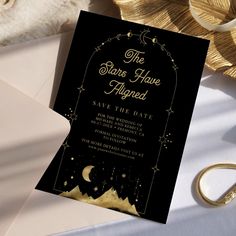 a black and gold save the date card on top of a white table cloth next to a pair of wedding rings