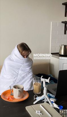 a woman wrapped in a blanket sitting at a table with a laptop and coffee mug