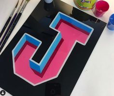 the letter j is painted pink and blue on a black background with paintbrushes next to it