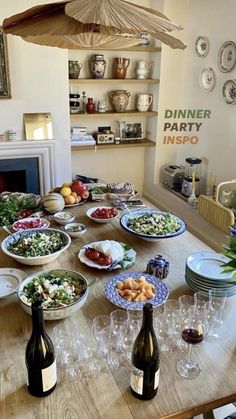 a wooden table topped with lots of plates and bowls filled with food next to bottles of wine