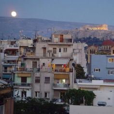 the full moon is setting over an urban area with buildings and hills in the background