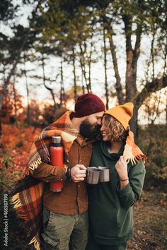 a man and woman standing next to each other in the woods holding coffee mugs