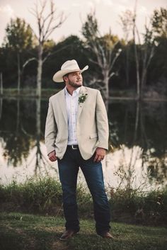 a man standing in front of a lake wearing a cowboy hat and suit with his hands in his pockets