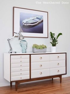 a white dresser topped with lots of drawers next to a painting on the wall above it