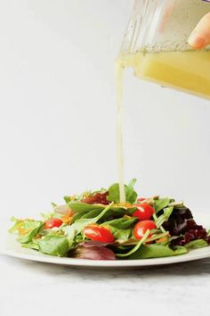 someone pouring dressing onto a salad with lettuce, tomatoes and other vegetables on a white plate