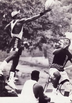 two men are playing basketball in the park while others watch from the sidelines and look on