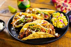 three tacos on a black plate with corn, salsa, and cole slaw