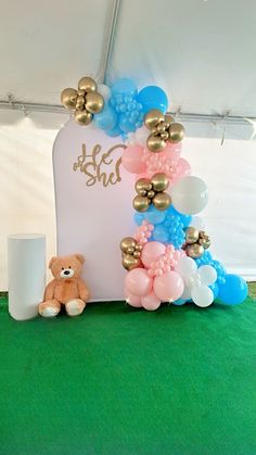 a teddy bear sitting in front of a sign with balloons on it's side