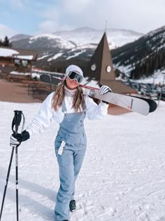 a woman in overalls and goggles holding a snowboard on her shoulder while standing in the snow