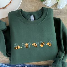 a green sweatshirt with bees embroidered on the front and back, sitting next to a pair of jeans