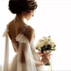 a woman in a white dress holding a bouquet and looking off into the distance with her back to the camera