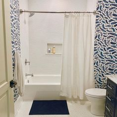 a white bath tub sitting next to a toilet under a bathroom window covered in blue and white wallpaper