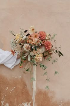 a bridal bouquet with peach and orange flowers is held against a wall by someone's hand