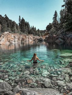 a woman swimming in the middle of a lake surrounded by rocks and pine trees with clear blue water