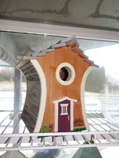 a birdhouse with a purple door and brown roof sitting on a shelf in front of a window