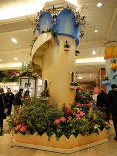 people are walking around in an indoor shopping mall with plants and flowers on display at the entrance