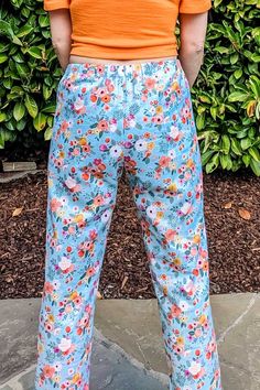 a woman standing in front of a bush with her back to the camera, wearing blue floral pants and an orange shirt