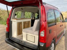 the back end of a red van with its doors open on a dirt road next to grass and trees