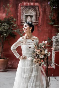 a woman standing in front of a red building wearing a white dress and holding a bouquet