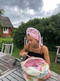 a woman sitting at a picnic table with a pink towel on her head and scissors in her hand