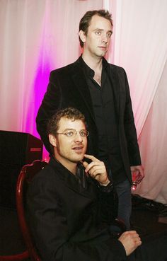 two men sitting next to each other in front of a pink curtain and one holding a cell phone