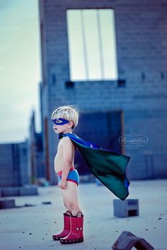 a young boy dressed as a super hero standing in front of a building with his cape around his neck