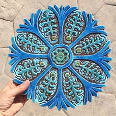 a hand holding up a blue and brown decorative object on a stone floor with rocks in the background