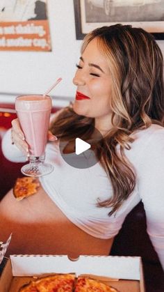 a woman sitting at a table with a box of pizza and a drink in her hand