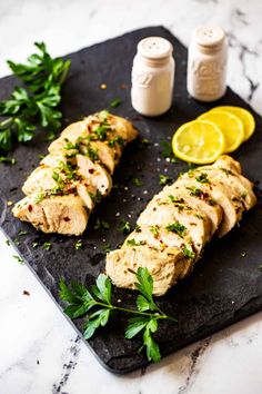 chicken breast with herbs and lemon on a black cutting board next to some condiments