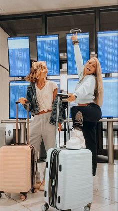 two women are standing in an airport with their luggage and pointing at the screen behind them