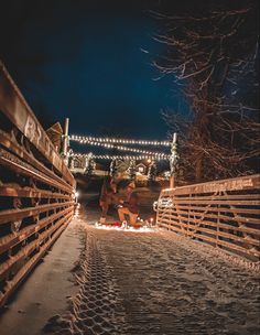 two people are standing in the middle of a snowy path with lights strung over them