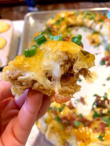 a person holding up a piece of food with cheese and green onions on it in front of a casserole dish