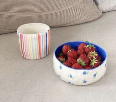 a bowl of strawberries sitting next to a striped cup on the floor in front of a pillow