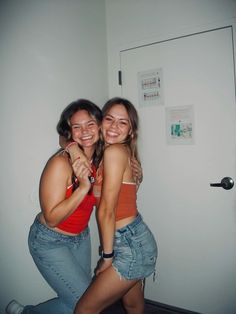 two young women hugging each other in front of a white wall and door, both smiling