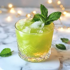 a green drink with ice and mint garnish on a marble counter top next to christmas lights