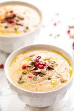 two white bowls filled with soup on top of a marble counter topped with pumpkin seeds and cranberries