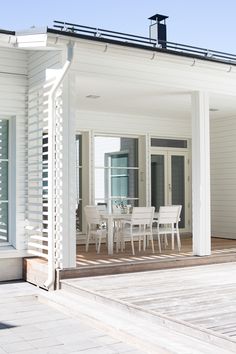 an outdoor dining area with white chairs and table on wooden deck next to large windows