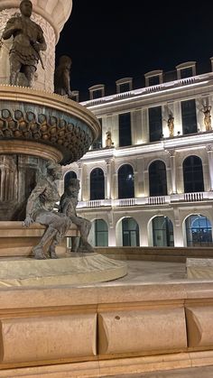 a fountain in front of a building with statues on the sides and lights at night