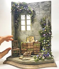 a miniature house made out of wood and flowers on the outside, in front of a person's hand