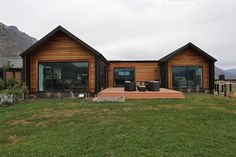 a wooden house with two large windows sitting on top of a lush green field next to mountains