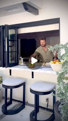 a man preparing food on top of a counter next to two stools in front of an open window