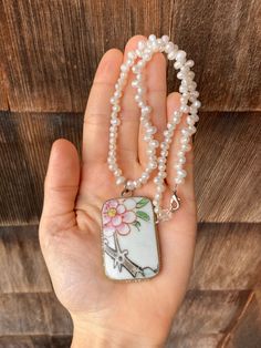 This pretty, Asian style necklace has a rectangular, ceramic pendant with a painted pale pink cherry blossom and decorative silver toned border. The strand is lined with what appear to be real pearls and there is a lobster claw clasp at the top. The pendant measures 2 inches long by 1 1/8 inches wide and the strand is 17 inches long from end to end. The pendant shows minor wear but overall the necklace is in good condition. White Rectangular Pendant Necklace For Gift, White Necklace With Rectangular Pendant For Gift, Handmade White Rectangular Pendant Jewelry, White Square Necklace For Gift, Hand Painted Rectangular Jewelry As Gift, Elegant White Hand Painted Necklaces, Vintage Pearl Necklace, Cherry Blossom Painting, Pink Cherry Blossom